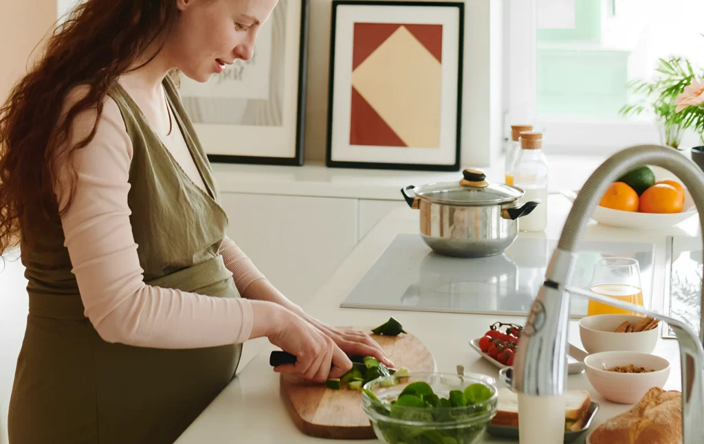 En la imagen podemos ver a una mujer preparando una deliciosa y saludable comida para su embarazo. Se encuentra en su cocina, rodeada de diferentes ingredientes y utensilios de cocina. Con una sonrisa en su rostro, la mujer muestra cómo la alimentación es una parte importante del cuidado durante el embarazo. Su elección de alimentos frescos y saludables es un gran ejemplo de cómo una buena nutrición puede mejorar tanto la salud de la madre como la del bebé en crecimiento